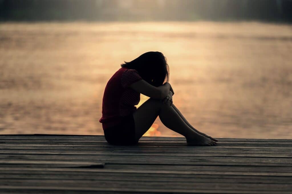Woman sitting on decking by water with head in arms resting on legs